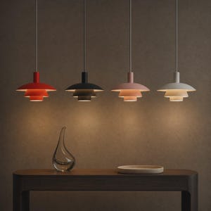 May include: Four pendant lights hang in a row, each featuring a tiered, multi-layered shade design. The colors from left to right are bright red, matte black, soft dusty pink, and off-white. Each lamp is suspended by a thin, vertical cord.
