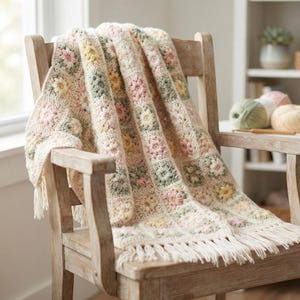 Puede incluir: Una manta de ganchillo con un patrón repetitivo de cuadrados de la abuela con centros florales en tonos suaves de rosa, amarillo y verde salvia sobre un fondo crema, terminada con un fleco anudado a lo largo del borde inferior del tejido.