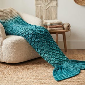 May include: A crochet mermaid tail blanket featuring a color gradient that transitions from a light beige at the top to a deep teal at the bottom. The blanket has a textured scale pattern throughout the body and a ribbed fin at the end.