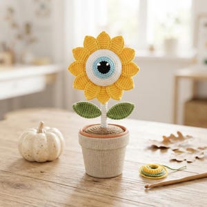 May include: A crocheted sunflower with a large central eye design featuring a black pupil, light blue iris, and white sclera. The flower has yellow petals, a white stem, and two green leaves, all potted in a beige fabric-covered container.