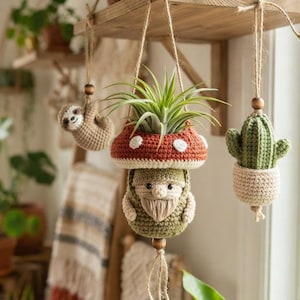 Op de afbeelding: Drie gehaakte hangende ornamenten aan touwtjes. Het middelste stuk is een kabouter met een rode paddenstoelhoed met witte stippen, die een groene luchtplant vasthoudt. Links hangt een kleine beige luiaard en rechts een groene cactus.