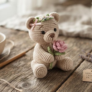 May include: A beige crocheted teddy bear with a flower crown and a pink rose in its paws. The bear has dark eyes and a brown stitched nose. A small tag nearby reads HANDMADE CROCHET PATTERN with a heart symbol. A ball of yarn and a hook are visible.