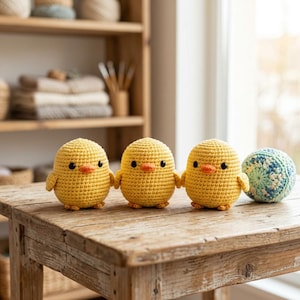 Op de afbeelding: Drie kleine, ronde, gele gehaakte kuikentjes met oranje snavels en zwarte kraaloogjes, op een rij geplaatst naast een kleine, ronde, blauw-witte gehaakte bal op een houten oppervlak. Elk kuikentje heeft een getextureerd, garenachtig uiterlijk.