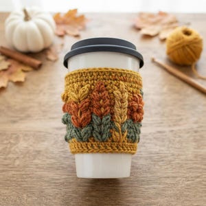 Könnte beinhalten: Ein weißer, wiederverwendbarer Kaffeebecher mit einer strukturierten Häkelmanschette. Die Manschette zeigt ein Muster aus erhabenen, gestickten Blättern und Weizenähren in Senfgelb, Rostorange und Salbeigrün, abgeschlossen mit einem schwarzen Kunststoffdeckel.