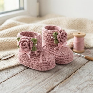 Könnte beinhalten: Ein Paar altrosa gehäkelte Babyschuhe mit einer dekorativen Rose, grünen Blättern und kleinen weißen Perlen an der Seite. Jeder Schuh ist mit einer passenden rosa Schleife vorne und einem gehäkelten Schnürverschluss am Knöchel versehen.