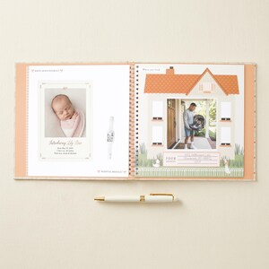 May include: A spiral-bound baby memory book open to two pages. The left page features a birth announcement for Lily Rose with a photo of a newborn, text details, and a hospital bracelet. The right page shows a house illustration with a photo of a person holding a baby carrier and an address section. A white and gold pen lies horizontally below the book.