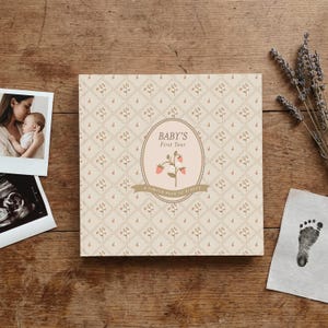 May include: A square baby memory book with a cream-colored cover featuring a repeating diamond pattern with small floral sprigs. The center oval label displays the text "BABY'S First Year" and "A SIMPLE BOOK OF FIRSTS" with a strawberry illustration.