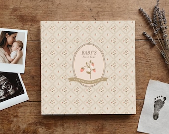 May include: A square baby memory book with a cream-colored cover featuring a repeating diamond pattern with small floral sprigs. The center oval label displays the text "BABY'S First Year" and "A SIMPLE BOOK OF FIRSTS" with a strawberry illustration.