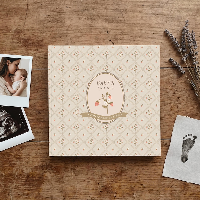 May include: A square baby memory book with a cream-colored cover featuring a repeating diamond pattern with small floral sprigs. The center oval label displays the text "BABY'S First Year" and "A SIMPLE BOOK OF FIRSTS" with a strawberry illustration.