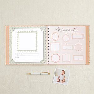 May include: An open spiral-bound memory book with a light peach cover. The left page features a scalloped photo frame and lines for notes. The right page is titled "These are a few of your FAVORITE THINGS" with various frames. A white pen and a baby photo are below.