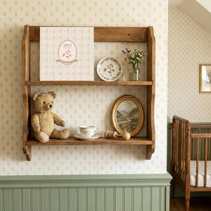 May include: A two-tiered wooden wall shelf holding a Baby's First Year book, a small plate with strawberry illustrations, a glass vase with wildflowers, a teddy bear, a floral teacup and saucer, a wooden rattle, and an oval landscape painting.
