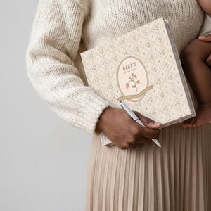 May include: A square baby memory book with a cream-colored cover featuring a repeating floral pattern. The center oval label displays the text "BABY'S First Year" above an illustration of a pink flower with green leaves and a tan ribbon banner.