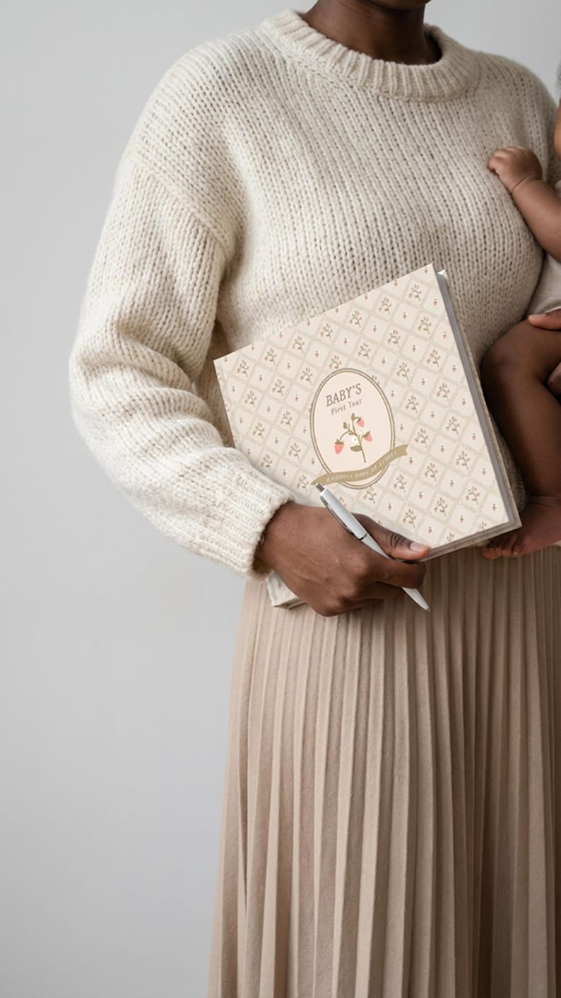 May include: A square baby memory book with a cream-colored cover featuring a repeating floral pattern. The center oval label displays the text "BABY'S First Year" above an illustration of a pink flower with green leaves and a tan ribbon banner.