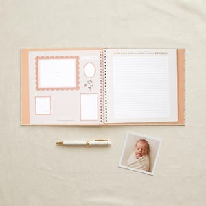 May include: A spiral-bound baby memory book with a light peach cover, featuring internal pages with a grid pattern, scalloped photo frames, and lined writing space. Includes a white pen and a square photograph of a swaddled infant.