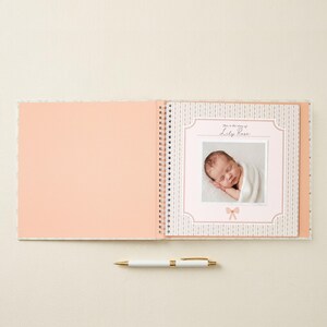 May include: A spiral-bound baby memory book with a solid peach left page and a patterned right page featuring a central frame. The frame contains a photograph of a sleeping infant wrapped in a white blanket, with the text "This is the story of Lily Rose" and a small bow icon. A white and gold pen lies horizontally below the book.