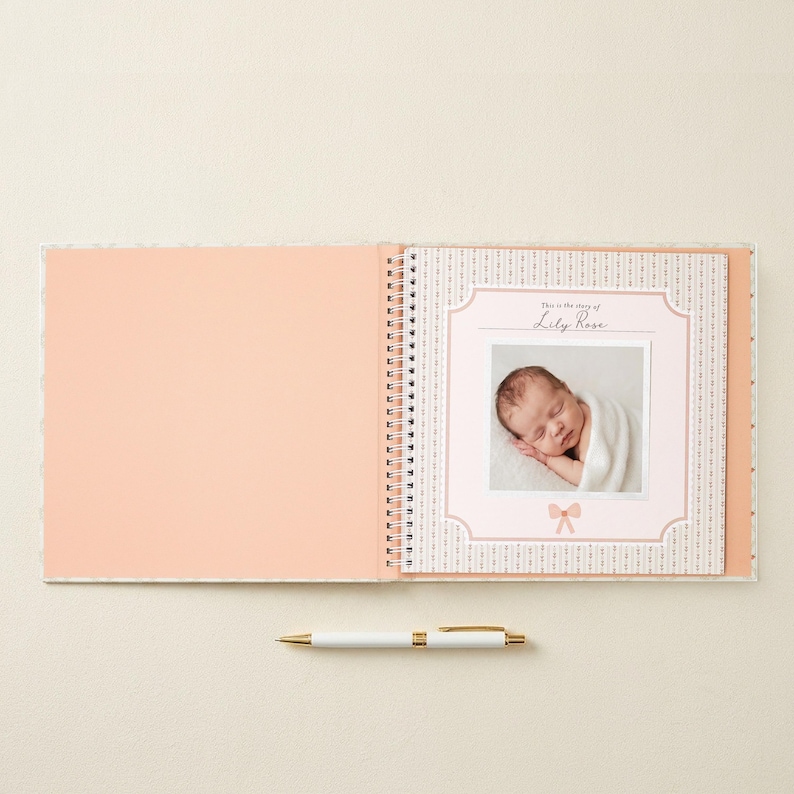 May include: A spiral-bound baby memory book with a solid peach left page and a patterned right page featuring a central frame. The frame contains a photograph of a sleeping infant wrapped in a white blanket, with the text "This is the story of Lily Rose" and a small bow icon. A white and gold pen lies horizontally below the book.