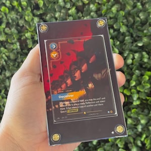 May include: A rectangular acrylic display case holding a Deceiver trading card with a red and black illustration of multiple faces. The case features four gold-colored circular screws at the corners, securing the card within the clear protective frame.