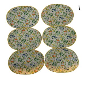 May include: A set of six oval placemats featuring a white background with a repeating pattern of green trees, orange fruits, yellow lemons, and blue berries. Each placemat is finished with a bright yellow fabric border around the entire edge.