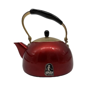 May include: A metallic red tea kettle with a brass-toned handle and a black handle grip. The kettle features a black knob on the lid and a small black label on the front with a white graphic of a person and the text ILOVAN TIL OG KAN.
