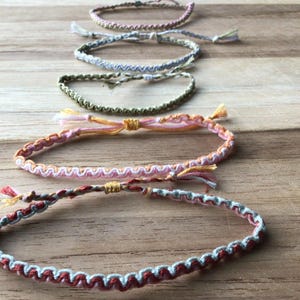 May include: A collection of five handmade woven bracelets arranged in a line. The bracelets feature various colour combinations including pink, white, grey, olive green, and orange, with intricate knotted patterns and loose thread ends at the closures.