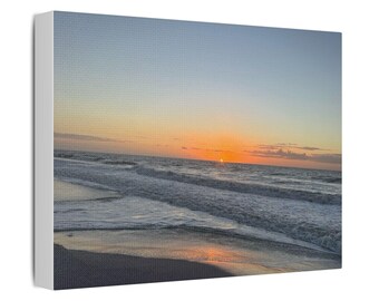 Könnte beinhalten: Ein rechteckiger Leinwanddruck mit einem Sonnenuntergang über dem Ozean. Am Horizont versinkt eine leuchtend orangefarbene Sonne im Wasser, während der Himmel in sanfte Blau- und Orangetöne getaucht ist. Sanfte Wellen rollen an den Sandstrand.