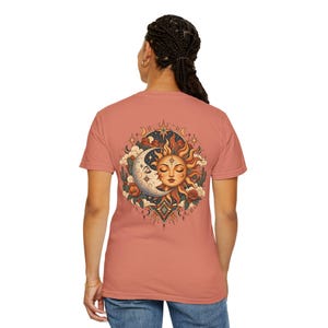 May include: A dusty rose short-sleeve t-shirt featuring a large circular graphic on the back. The design depicts a stylized sun and crescent moon with closed eyes, surrounded by clouds, stars, red flowers, and cacti in a warm color palette.