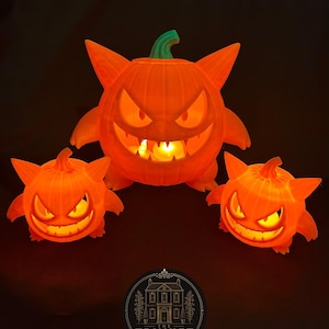 May include: Three orange, glowing, pumpkin-shaped figures with pointed ears and wide, toothy grins. The central figure is larger than the two smaller ones on either side. Each features a green stem on top and internal lighting. A circular logo reads THE TRINKET HOUSE.