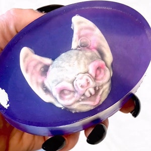 May include: A purple and white oval-shaped soap bar with a detailed bat design. The bat has a white face with pink ears and a toothy grin.
