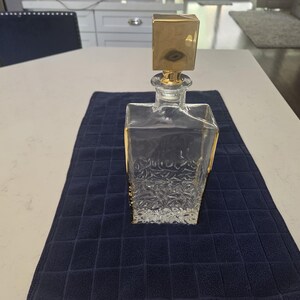 May include: A clear glass decanter with a square base and a textured, crystalline pattern on the lower half. The decanter features a smooth, transparent neck and is topped with a polished, square-shaped gold-toned stopper that fits securely.