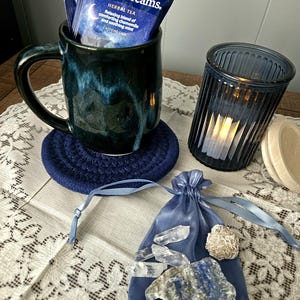 May include: A dark blue ceramic mug containing Bigelow Sweet Dreams herbal tea packets, resting on a navy blue rope coaster. Beside it sits a ribbed blue glass candle holder and a light blue satin drawstring pouch holding several raw crystals.