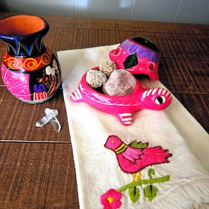 May include: A hand-painted black vase with orange, pink, and blue patterns, a pink turtle-shaped trinket dish with a matching lid containing three crystals, two clear quartz points, and a white cloth featuring an embroidered pink bird and flower.