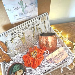 May include: A gift box containing an Arizona license plate, a hammered copper mug, a clear shot glass inside an orange velvet scrunchie, a small bowl with green stones, and two cards with text including The Cactus in the Clouds and The Copper Queen.