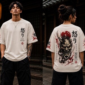 Könnte beinhalten: Ein cremefarbenes Kurzarm-T-Shirt mit einem Grafikdruck einer japanischen Oni-Maske auf der Rückseite, roten Spritzern und dem Text "ONI'S WRATH" sowie japanischen Schriftzeichen. Die Vorderseite zeigt kleineren japanischen Text und rote Details.