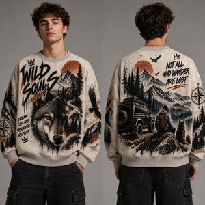 Op de afbeelding: Een beige sweater met ronde hals en een grafische print van een wolf, bergen en bos op de voorkant met de tekst "WILD SOULS" en "DREAM EXPLORE DISCOVER REPEAT". De achterkant toont een voertuig, een persoon en "NOT ALL WHO WANDER ARE LOST".
