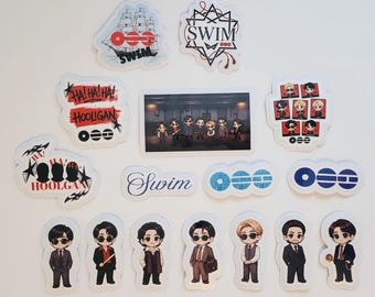 Puede incluir: Una colección de pegatinas con varios diseños, incluyendo un barco, el texto SWIM, HOOLIGAN y HA! HA! HA!, junto con una pegatina rectangular que muestra un grupo de figuras y varias pegatinas individuales de personajes estilo chibi en traje.