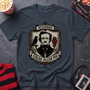 May include: A dark gray t-shirt featuring a graphic illustration of Edgar Allan Poe centered within a decorative shield. The design includes a black raven, a red anatomical heart, and the text NEVERMORE and EDGAR ALLAN POE in a banner.