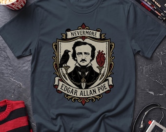 May include: A dark gray t-shirt featuring a graphic illustration of Edgar Allan Poe centered within a decorative shield. The design includes a black raven, a red anatomical heart, and the text NEVERMORE and EDGAR ALLAN POE in a banner.