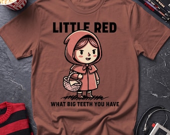 May include: A brown short-sleeved t-shirt featuring a cartoon illustration of a character in a red hooded cloak holding a basket. The text "LITTLE RED" appears above the character, and "WHAT BIG TEETH YOU HAVE" is printed below the illustration.