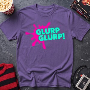 May include: A purple short-sleeved t-shirt featuring a bright pink ink-blot graphic on the chest. The text GLURP GLURP! is printed in a bold, light green sans-serif font across the center of the pink shape, creating a high-contrast design.