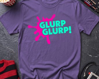 May include: A purple short-sleeved t-shirt featuring a bright pink ink-blot graphic on the chest. The text GLURP GLURP! is printed in a bold, light green sans-serif font across the center of the pink shape, creating a high-contrast design.
