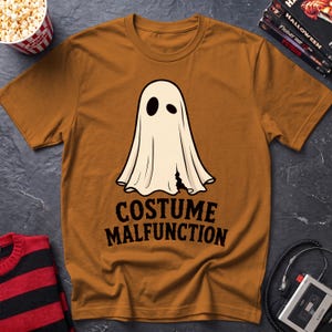 May include: A mustard yellow short-sleeve t-shirt featuring a graphic of a white ghost with two black eye holes and a small tear at the bottom. Below the ghost, the text "COSTUME MALFUNCTION" is printed in bold, black, capital letters.
