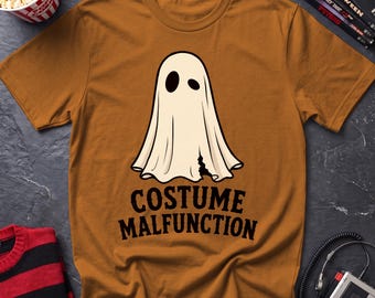 May include: A mustard yellow short-sleeve t-shirt featuring a graphic of a white ghost with two black eye holes and a small tear at the bottom. Below the ghost, the text "COSTUME MALFUNCTION" is printed in bold, black, capital letters.