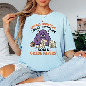 May include: A light blue t-shirt featuring a graphic of a purple one-eyed monster sitting at a desk, writing on papers with a mug nearby. The text reads: NOT ALL MONSTERS LIVE UNDER THE BED SOME GRADE PAPERS. The design uses orange and black ink.