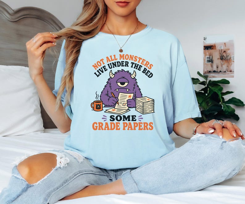 May include: A light blue t-shirt featuring a graphic of a purple one-eyed monster sitting at a desk, writing on papers with a mug nearby. The text reads: NOT ALL MONSTERS LIVE UNDER THE BED SOME GRADE PAPERS. The design uses orange and black ink.
