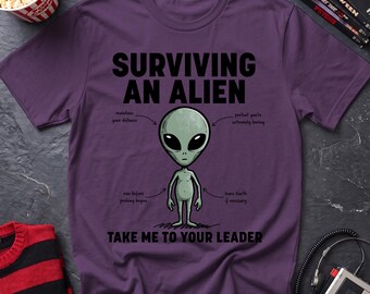 May include: A purple t-shirt featuring a graphic of a grey alien in the center. The text reads "SURVIVING AN ALIEN" at the top and "TAKE ME TO YOUR LEADER" at the bottom. Arrows point to the alien with instructions: "maintain your distance", "pretend you're extremely boring", "run before probing begins", and "leave Earth if necessary".