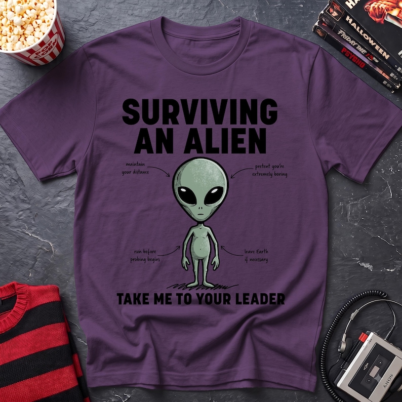 May include: A purple t-shirt featuring a graphic of a grey alien in the center. The text reads "SURVIVING AN ALIEN" at the top and "TAKE ME TO YOUR LEADER" at the bottom. Arrows point to the alien with instructions: "maintain your distance", "pretend you're extremely boring", "run before probing begins", and "leave Earth if necessary".
