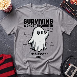 May include: A grey t-shirt featuring a graphic of a white ghost with the text "SURVIVING A GHOST ENCOUNTER" and "BOO!". The design includes arrows pointing to the ghost with labels reading "eye holes limit visibility", "offer to do his laundry", and "don't trip on the sheet", plus "sheet snags easily".