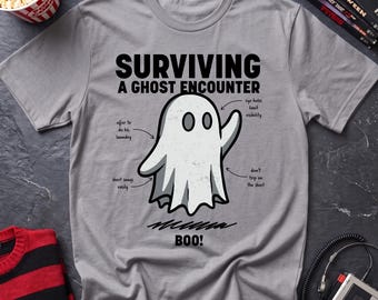 May include: A grey t-shirt featuring a graphic of a white ghost with the text "SURVIVING A GHOST ENCOUNTER" and "BOO!". The design includes arrows pointing to the ghost with labels reading "eye holes limit visibility", "offer to do his laundry", and "don't trip on the sheet", plus "sheet snags easily".