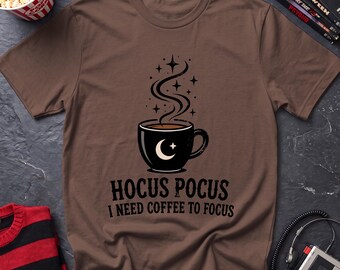 May include: A brown short-sleeve t-shirt featuring a black coffee mug graphic with a white crescent moon and star. Above the mug is rising steam with small stars. Below the graphic is the black text: HOCUS POCUS I NEED COFFEE TO FOCUS.