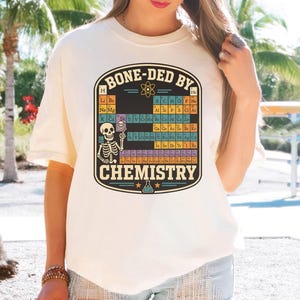 May include: A cream-colored t-shirt featuring a graphic print of a periodic table of elements with a skeleton pointing at a skull icon. The text BONE-DED BY CHEMISTRY is printed in a bold, dark font above and below the colorful table design.